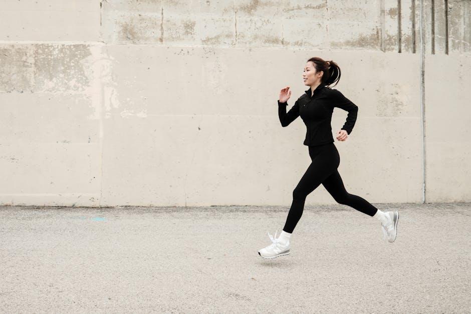 Woman in black athletic outfit running outdoors against a concrete wall, daytime.