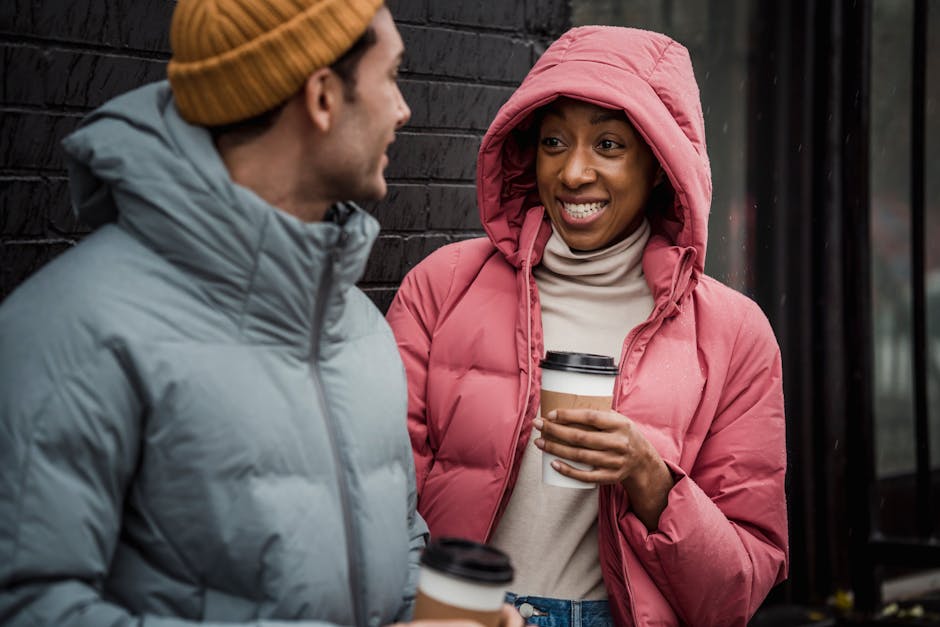 Cheerful multiracial couple in warm clothes with tasty takeaway hot coffee looking at each other while standing near building on street