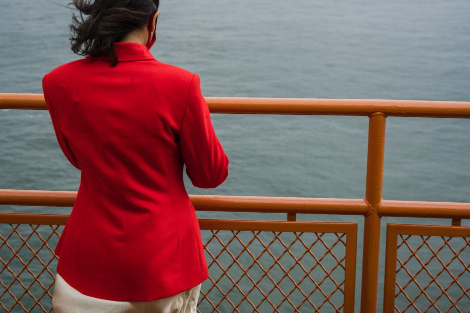 Back view of a woman in a red blazer leaning on a metal fence by the water.