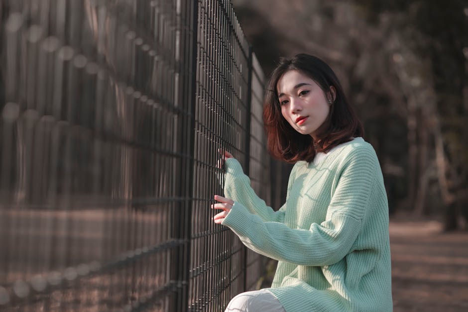 Elegant woman in a knitted mint sweater poses by a fence outdoors during daytime.