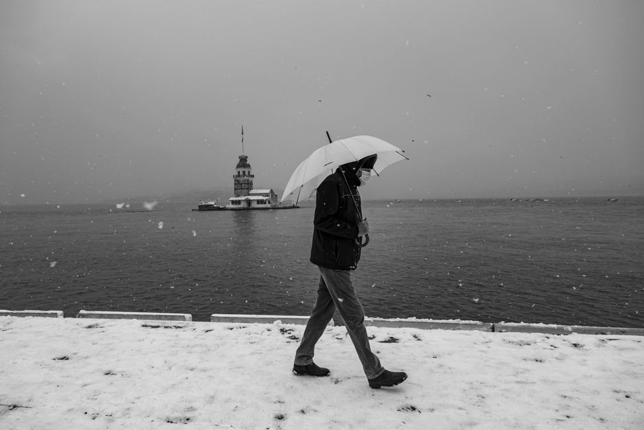 A person walks by a snowy river holding an umbrella with Maiden's Tower in the background.