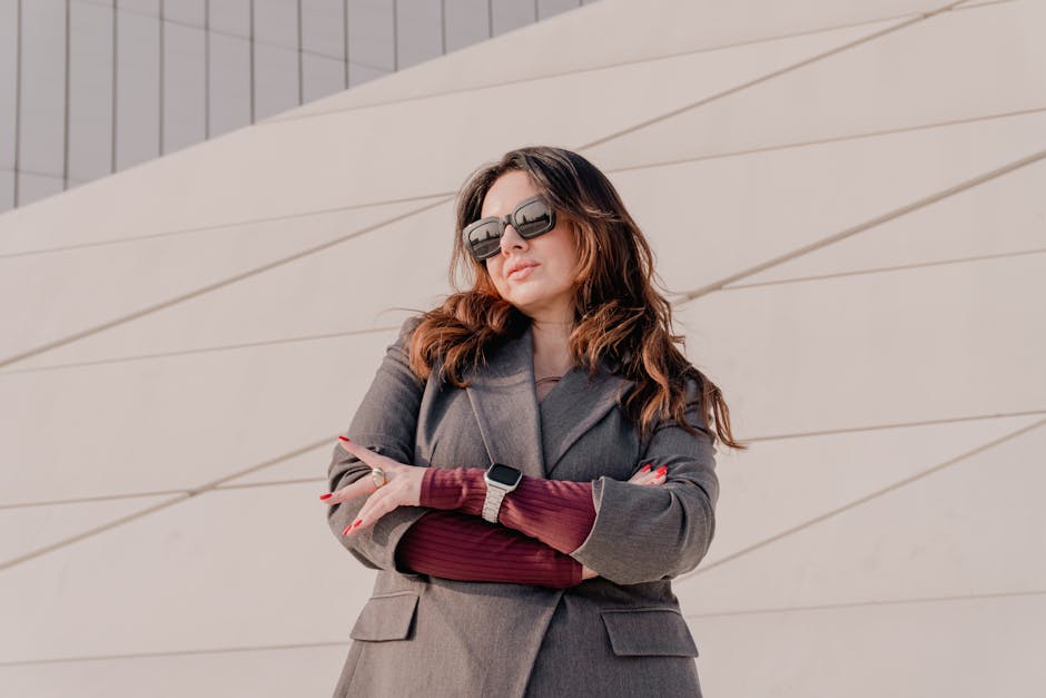 Fashionable woman posing confidently in sunglasses and gray coat against modern architecture.