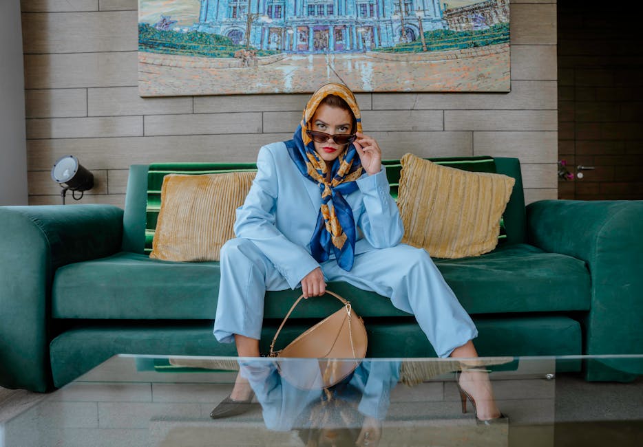 Fashionable woman in a blue suit with a scarf indoors, seated on a green couch with artwork backdrop.