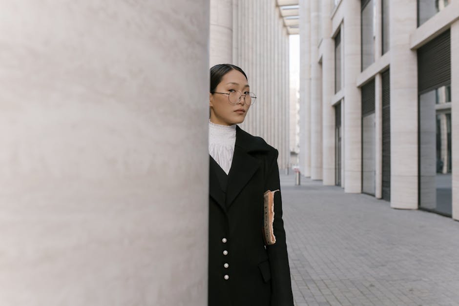 Asian businesswoman in glasses and coat standing in modern urban environment.