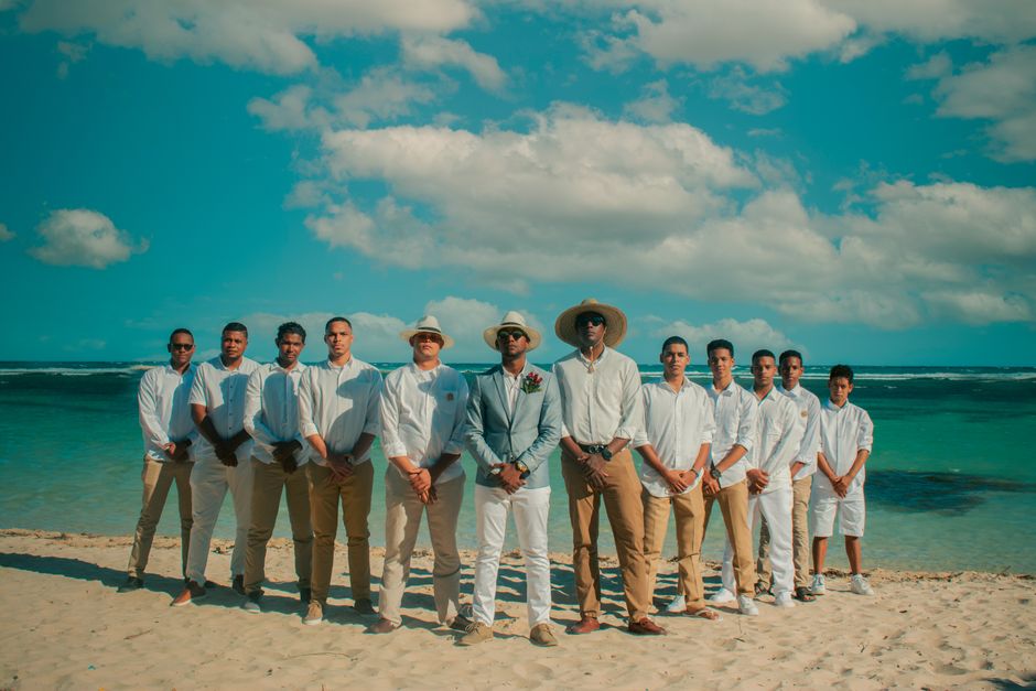 A group of groomsmen with the groom on a beach, wearing light attire under a sunny sky.