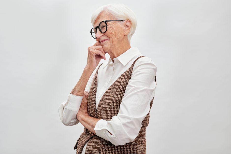 Stylish elderly woman with white hair and eyeglasses, pondering happily indoors.