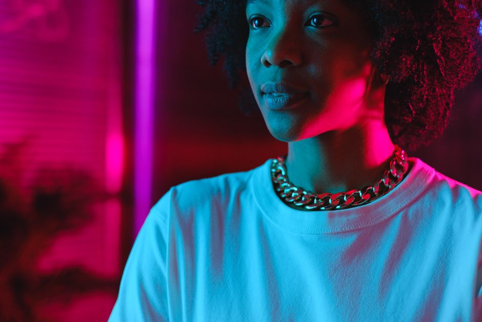 Crop positive African American female in white shirt standing in pink neon lights in dark studio and looking away