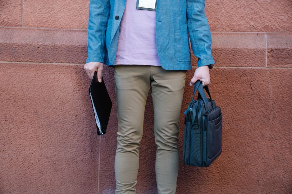 A man standing against a wall holding a briefcase and documents, dressed in casual business attire.