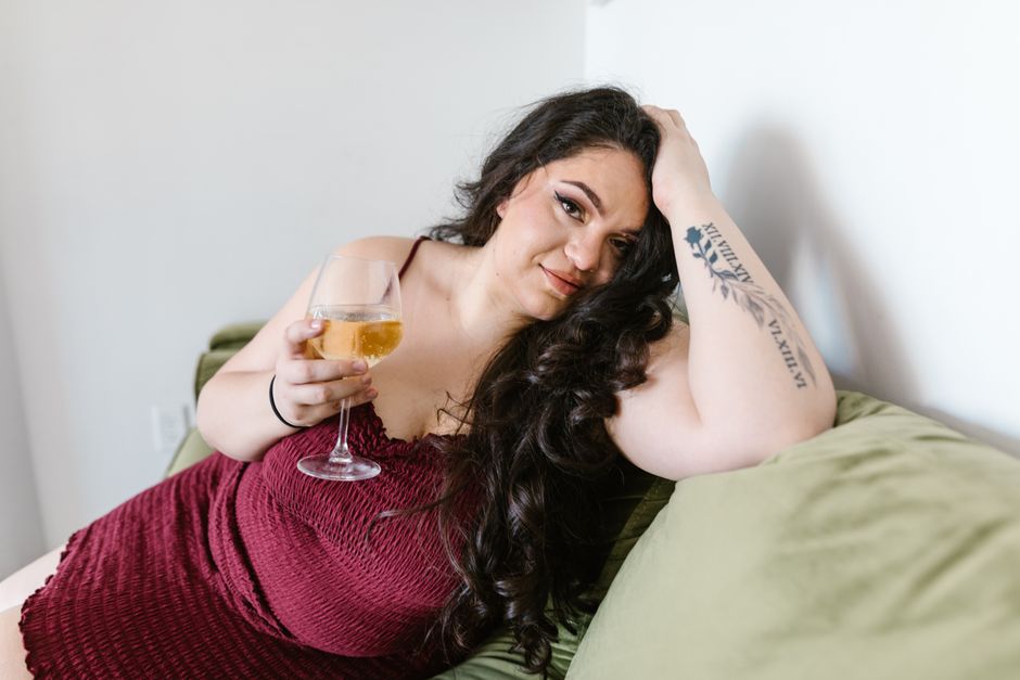 Curvy woman in maroon dress relaxing on a green sofa, enjoying a glass of wine.