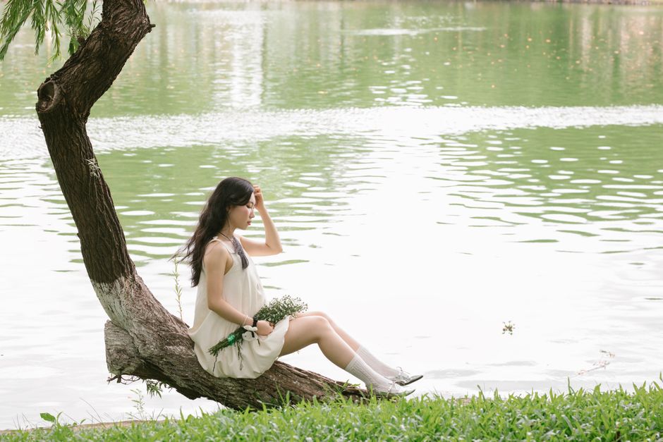 Young woman in a white dress relaxing by a tranquil lake with a bouquet of flowers.