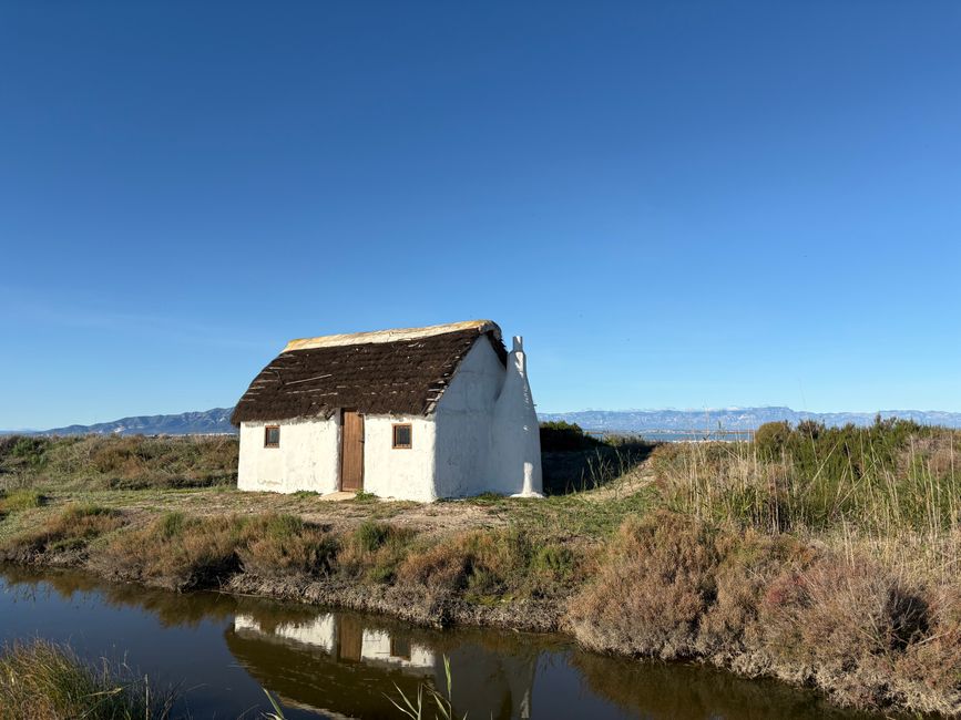 A rustic white house with a thatched roof by a peaceful stream under a clear blue sky.
