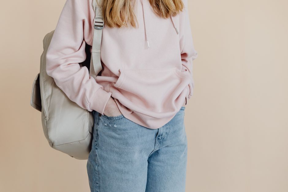 A teenager in a casual pink hoodie and jeans carrying a backpack, standing against a neutral background.