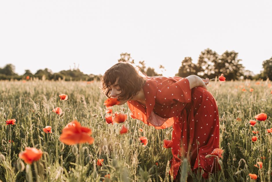 A woman in a red polka dot dress bends to smell vibrant poppies in a sunny field.