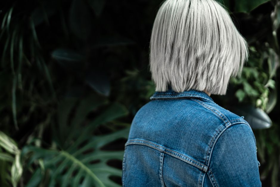 Woman with white hair in denim jacket surrounded by lush greenery outdoors.