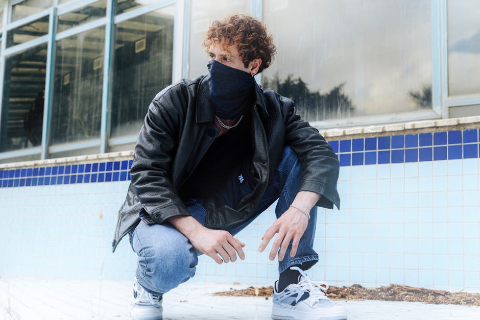 A young man crouches stylishly in an empty tiled swimming pool wearing a leather jacket and sneakers.