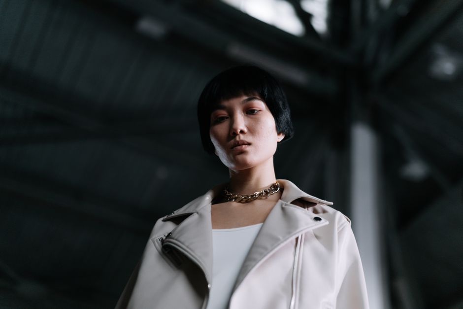 Low angle portrait of a woman with black hair and chain necklace, exuding confidence in an industrial setting.