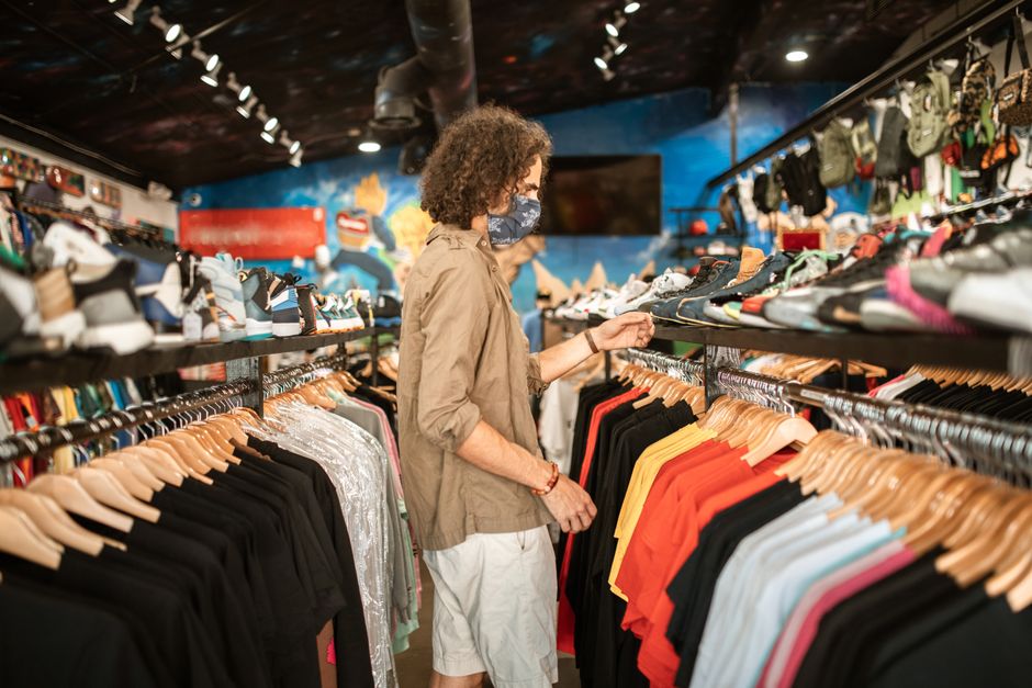 A fashionable young man shops for clothes in a trendy urban store, wearing a mask.