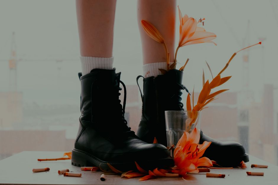 Close-up of black boots with vibrant orange lilies and scattered cigarette butts indoors.