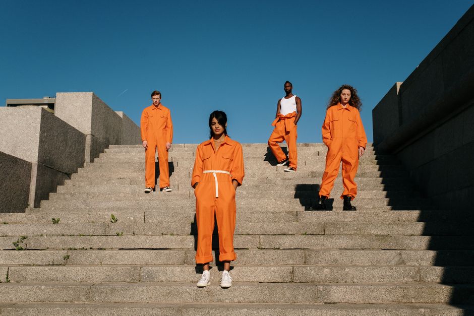 Four individuals in orange jumpsuits standing on outdoor concrete steps against a clear blue sky.